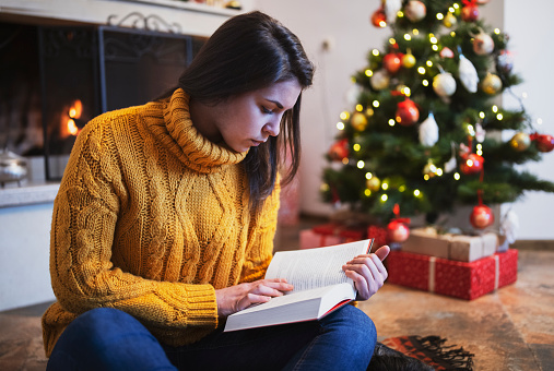 Young beautiful girl is reading a book next to the fireplace and the Christmas tree and the fireplace at the Christmas decorated background , Christmas holidays