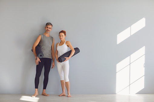 Yoga people. A man and a woman with yoga mats stand on a gray background in a bright classroom. Couple getting ready to practice yoga.