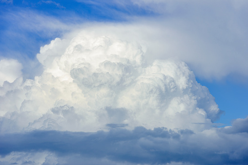 Fat cumulus clouds accumulate to build an enormous mountain of clouds, announcing heavy weather. A sky only, full-frame and close-up image of a tremendous cumulonimbus cloud. Some cirrus clouds appear high up in the blue sky and a band or roll cloud of stratocumuli extend low over the foreground. A sky only, full-frame and close-up image.