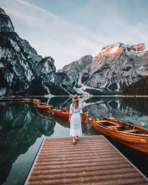 Rearview of young beautiful woman enjoy nature and summer walking on a wooden pier during sunrise and looking at the view with orange boats and mountain reflection at a beautiful turquoise water lake Lago di Braies, Trentino Alto Adige, South Tirol, European Alps, Italia, Europe