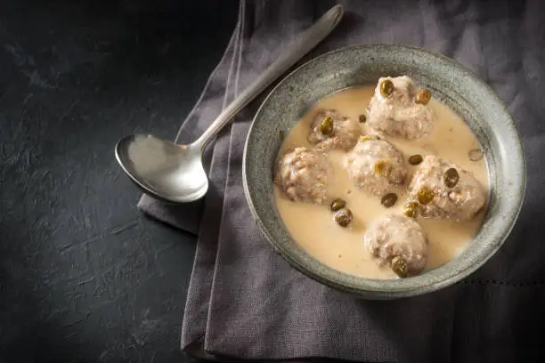 Boiled meatballs in a white bechamel sauce with capers, called Koenigsberger Klopse, traditional Polish and German dish in bowl on a dark gray background with copy space, view from above, selected focus