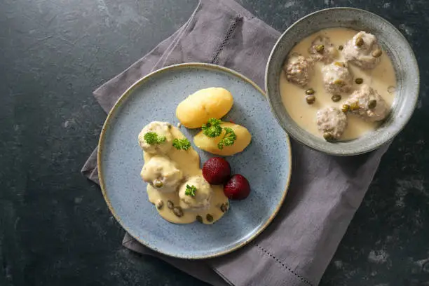 Koenigsberger Klopse or meatballs in white bechamel sauce with capers, potatoes and beetroot served in a bowl and on a gray blue plate, dark rustic background with copy space, high angle view from above
