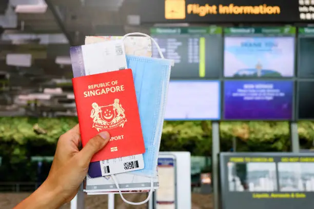 In airport: hand holding Singapore passport, boarding pass & mask, flight information board in background. Travel essential concept; reopening; post covid-19 coronavirus. Selective focus.
