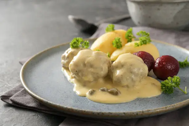 Koenigsberger Klopse or meatballs in a white bechamel sauce with capers, potatoes and beetroot served on a gray blue plate, traditional Polish and German dish, copy space, selected focus, narrow depth of field