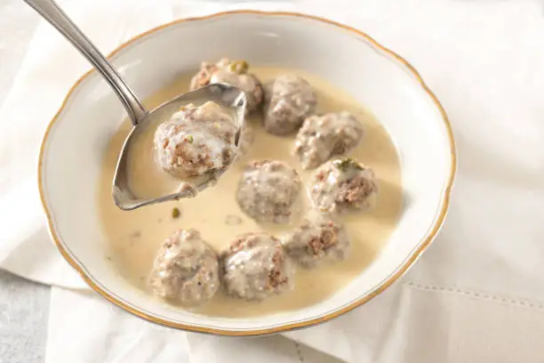 Ladle is liftiting a meatball from a bowl with koenigsberger klops, which are boiled beef balls in a white bechamel sauce with capers, traditional Polish and German dish, selected focus, narrow depth of field