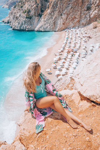 Young beautiful blonde woman in blue pareo and swimsuit sitting on a rock having fun and looking out of Kaputas Beach and sea view in Kaş, Kalkan, Mediterranean Sea, Turkiye.