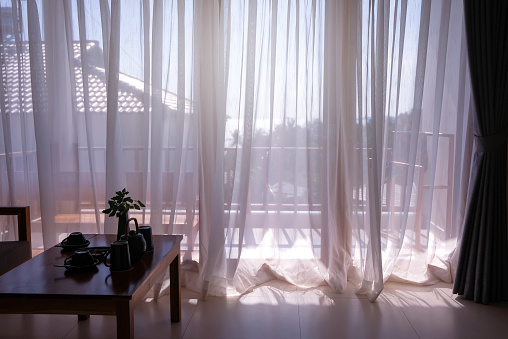 Room interior of beach front resort hotel, coffee table at the side. Partial view of sea seen through white translucent curtains at balcony windows. Soft sunlight; dreamy mood. Travel concept.