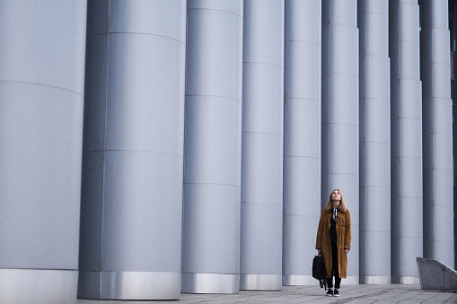 Young beautiful teenager model with long blonde hair wearing orange cape and black backpack going over huge columns in big city background. Lifestyle and fashion concept.