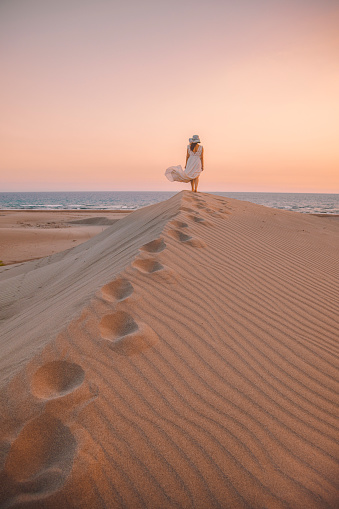 Rear view of young woman with white dress standing and walking on desert sand hill of Patara Beach watching beautiful colorful sunset in Kaş, Kalkan, Antalya, Türkiye
