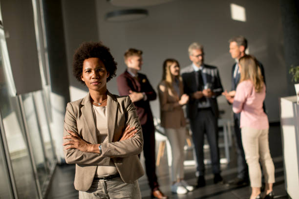 istock Professional black woman standing in office with folded arms and confident expression as other workers hold a meeting in background 1257550308