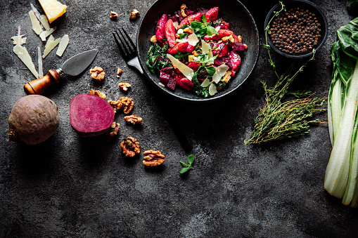 Top view of a freshly made beetroot pasta with ingredients around on a rustic table. Raw beetroot, black pepper, thyme, cheese, walnuts knife and and fork on black table.