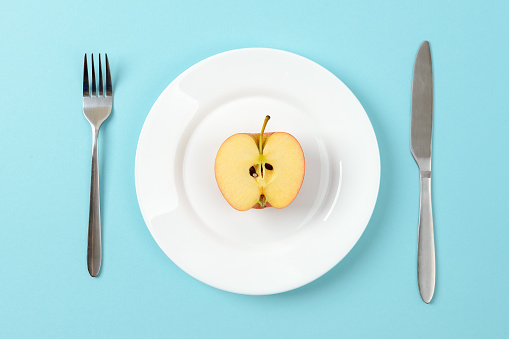 Half of ripe juicy apple on a white dinner plate, table knife and fork on a blue background. Concept of healthy eating with fiber and vitamins, vegetarianism and raw food diet. Top view.