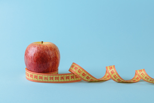 Ripe red apple and yellow measuring tape curles on a blue medical background. Concept of healthy eating, body weight control, weight loss and slimming. Copy space. Front view.