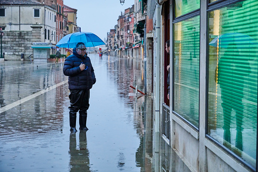 Venice, Italy - November 24, 2019: a man wearing rubber booth and an umbrella talking to the owner of a shop during the flood so called \