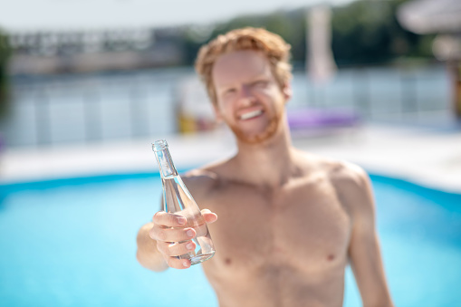 Life force. Bottle of refreshing drinking water in outstretched hand of young red-haired man near the pool