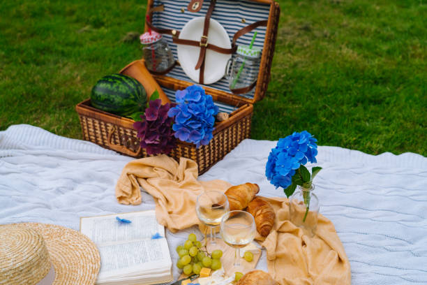 istock Summer picnic on sunny day with bread, fruit, bouquet hydrangea flowers, glasses wine, straw hat, book and ukulele. Picnic basket on grass with food and drink on white knit blanket. 1257591468