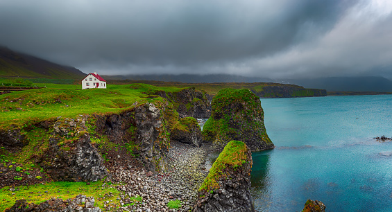 Lonely icelandic house with red roof on the sea coast with green grass meadow, rocks anf foggy sky. Natural Iceland travel landscape.