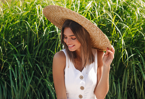 Happy stylish woman sitting in front of grass and looking while holding her hat