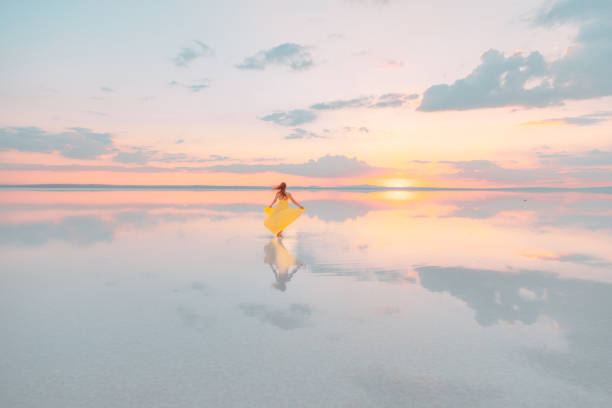 Rear view of young girl with yellow dress walking, posing, dancing and standing on Tuz Gölü with reflection on water and watching amazing sunset view and clouds in Lake Tuz located in the borders of Ankara, Aksaray and Konya, Türkiye