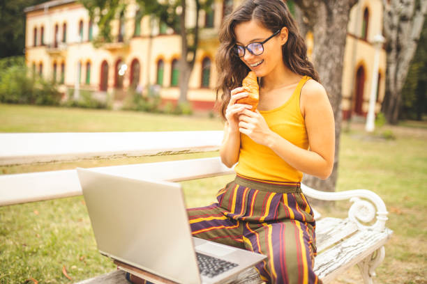 istock Woman using laptop, eating croissant and sitting on park bench 1257621629