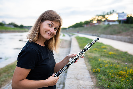 A woman is smiling and holding an oboe on a riverbank