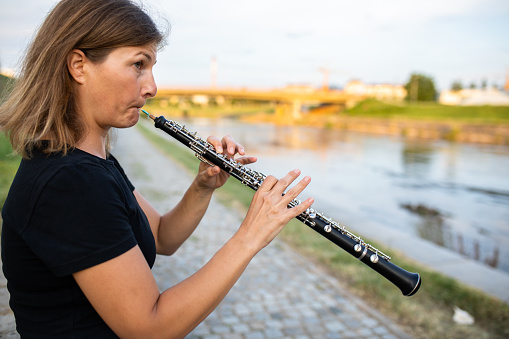 A woman is sitting on a stone wall by the river, playing the oboe