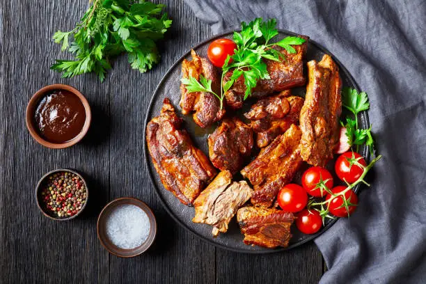 Restaurant slow-cooked pork ribs with bbq sauce, cherry tomato, and parsley on a black plate on a dark wooden background view from above, close-up
