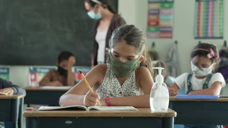 Group of students working at their desks in class all wearing protective face masks to avoid the transfer of germs after school reopened after COVID-19.