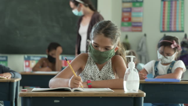 クラスの机で働く生徒のグループは、COVID-19の後に再開した後、放課後に細菌の移動を避けるために保護フェイスマスクを着用しています。