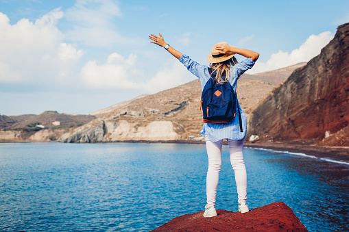 Summer vacation. Happy traveler woman feels free raising arms on rock on Red beach on Santorini island, Greece enjoying landscape.