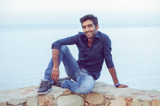 istock Man smiling looking at camera happy celebrating freedom while sitting on a concrete wall bridge above the sea. 1257646498