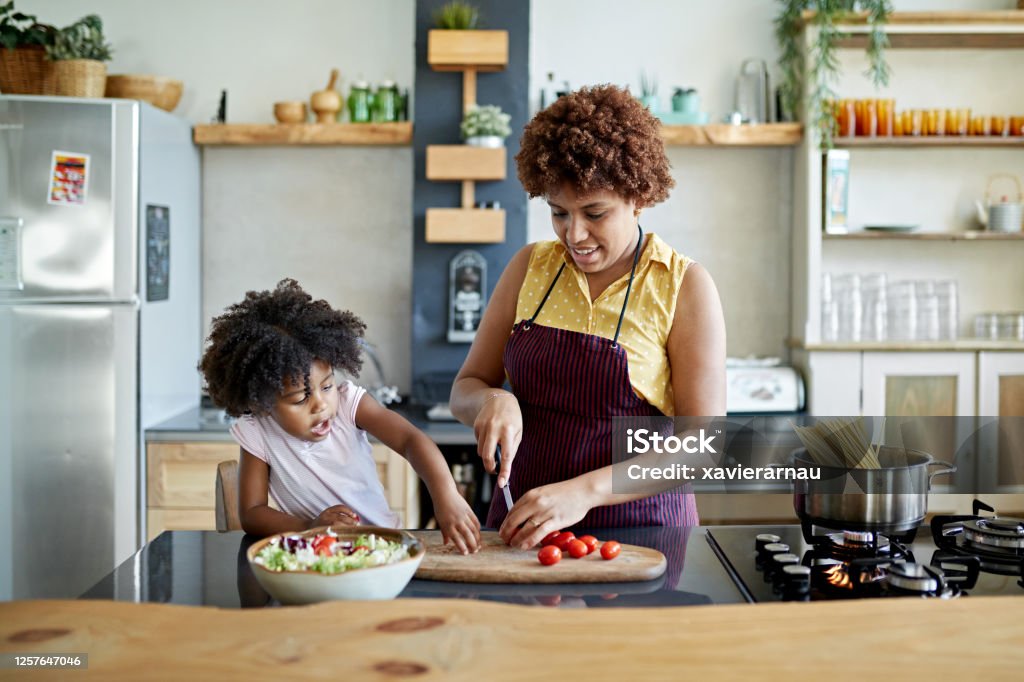 Afro-Caribbean Mother and Young Daughter Cooking Together Front view of 3 year old girl interacting with 26 year old mother in kitchen as she slices tomatoes on cutting board for salad and boils water for pasta. Healthy Eating Stock Photo