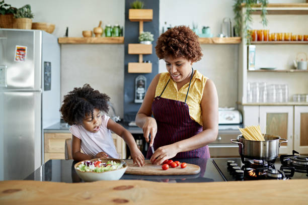 Afro-Caribbean Mother and Young Daughter Cooking Together Front view of 3 year old girl interacting with 26 year old mother in kitchen as she slices tomatoes on cutting board for salad and boils water for pasta. apron photos stock pictures, royalty-free photos & images