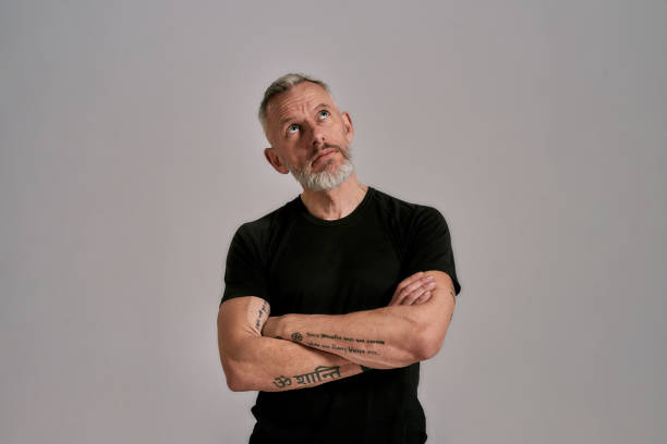 istock Middle aged man with crossed arms in black t shirt looking aside while posing in studio over grey background 1257649279
