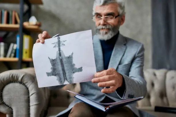 Photo of Mature psychologist holding picture with ink stain, Rorschach Inkblot during therapy session