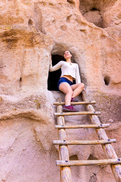 Happy woman climbing ladder on Main Loop trail in Bandelier National Monument in New Mexico during summer by canyon cliff cave Happy woman climbing ladder on Main Loop trail in Bandelier National Monument in New Mexico during summer by canyon cliff cave los alamos new mexico stock pictures, royalty-free photos & images
