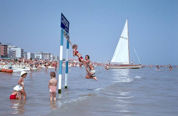 Rivazzurra, Rimini, Emilia-Romagna, Italy, 1966. On the beach in Rivazzurra. Old and young tumble in the water. Two young girls swing on the famous Campari swing. Also: hotels, tourists and a sailboat.