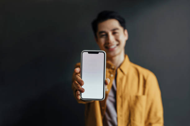 retrato de um homem asiático feliz de camisa amarela usando um celular/ distribuindo-o (tela em branco, espaço de cópia) - men blank holding showing - fotografias e filmes do acervo