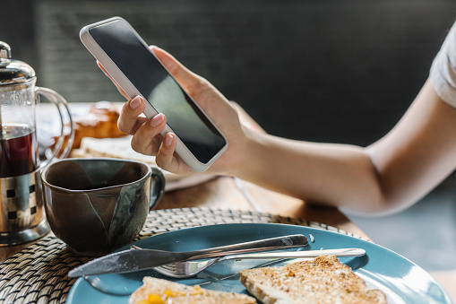 Anonymous woman holding a mobile phone while eating her breakfast.