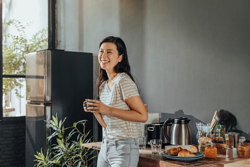Smiling young Asian woman wearing jeans and a t-shirt drinking tea for breakfast in the kitchen.