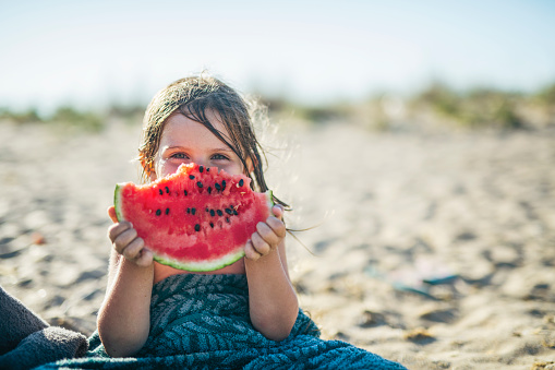 Little beautiful blue eyed girl is eating a watermelon at the beach and smiling