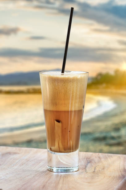 A greek cold coffee, freddo cappuccino placed on a wooden table over a beach background. A greek cold coffee, freddo cappuccino placed on a wooden table over a beach background at sunset. freddo cappuccino stock pictures, royalty-free photos & images