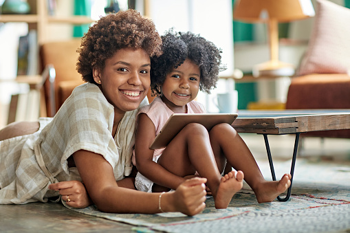 Front view of 26 year old Afro-Caribbean mother lying on living room rug with 3 year old daughter and smiling at camera as they use digital tablet together.