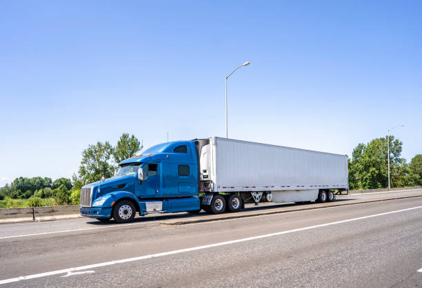 istock Blue big rig classic semi truck with high cab transporting load in refrigerator semi trailer running on the highway road entrance intersection 1257693031