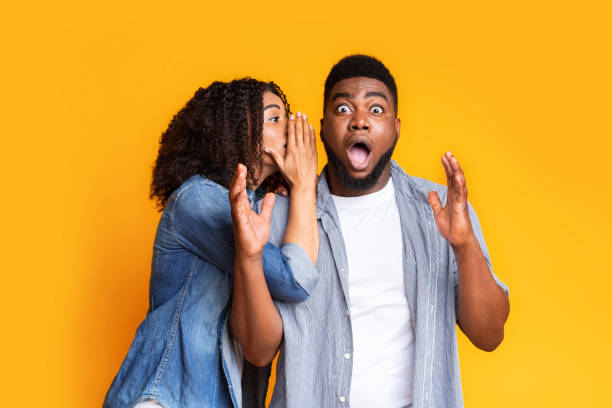 Shocking Offer. Black Girl Sharing Secret With Her Dazed Boyfriend Shocking Offer. Black Girl Sharing Secret With Her Dazed Boyfriend. Surprised Man Opens Mouth In Disbelief Over Yellow Studio Background slander stock pictures, royalty-free photos & images