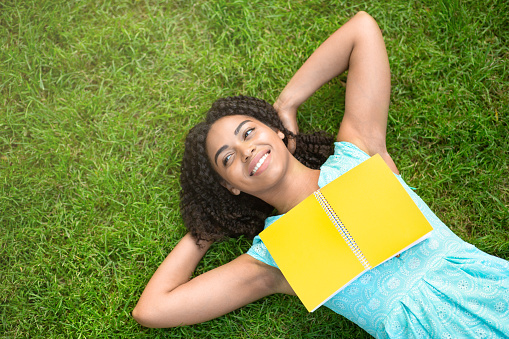 Overhead view of happy black girl lying on grass at park with open book, copy space
