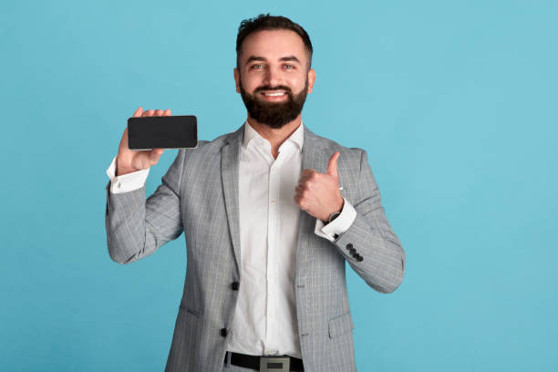 istock Happy office worker holding smartphone with blank screen and showing thumb up on blue background, space for design 1257706649