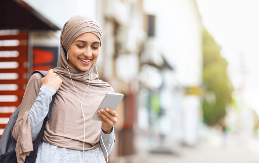 Stay always connected. Beautiful girl in hijab using earphones and newest smartphone, walking by street, empty space
