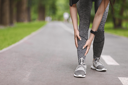 Young female runner suffering from shin splints while jogging in park, massaging painful area, having sport trauma, cropped image with copy space