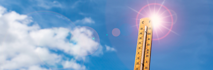 Wide image with wooden thermometer with high temperatures above 35 degrees Celsius or 95 degrees Fahrenheit. Measuring equipment against blue sky, clouds and sun with lens flare. Copy space. Weather banner.
